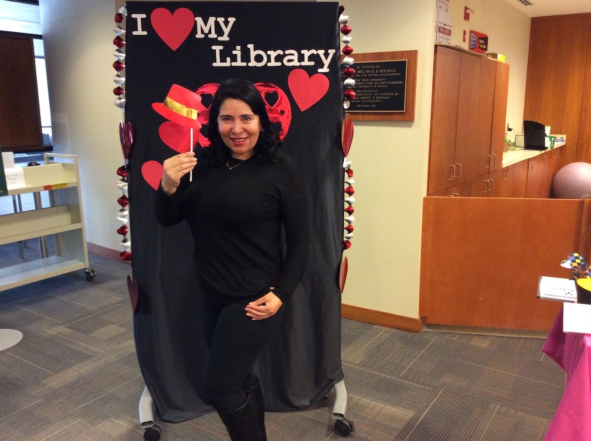 a smiling person wearing all black poses jauntily with a red hat prop on a stick in front of the I love my library backdrop